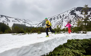Fotobehang Couple of cross country skiers man and woman with backpacks walking and exercising on ski in mountains near winter coniferous forest. Snow-covered mountain in full altitude on background © anatoliy_gleb