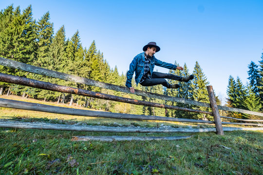 Young Male Farmer In A Denim Jacket And Cowboy Hat Jump Over The Fence