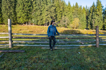 Young male farmer in a denim jacket and cowboy hat stand near the wooden  fence against forest 