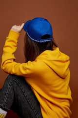 Back view shot of a brunette lady with long brown hair in a yellow hoodie and black jeans and blue baseball cap with white lettering. The girl is posing on the brown background. 