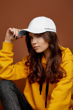 Half-turn shot of European lady with long brown hair in a yellow hoodie, black jeans and a white baseball cap with black lettering. The girl has black nails is posing on the brown background. 