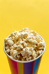 Popcorn in papercup. Multi coloured striped paper cup bucket with popcorn against a yellow background. Cinema, movies, streaming or entertainment concept. Vertical with copy space. 