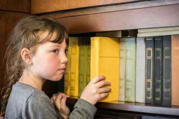 Serious little girl is taking a yellow book from the bookshelf. The child gets a textbook from the shelf of the old library. Children's educational concept