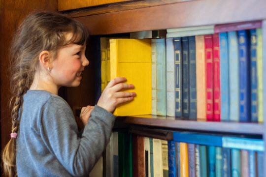 The Child Gets A Textbook With Wooden Shelf Of The Old Library. A Little Girl Is Taking A Yellow Book From A Bookshelf. Children's Educational Concept.