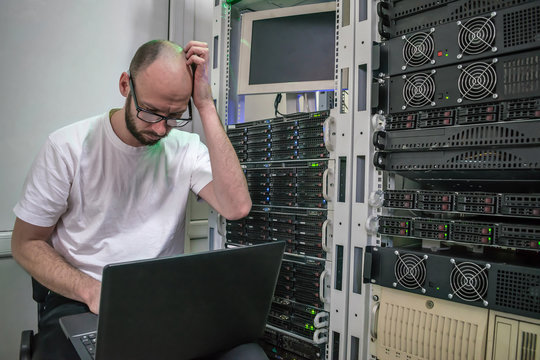 System administrator put his hand to his face while sitting in front of the computer. Man thinks about how to solve the problem in the server room. Programmer with a laptop thought in data center.
