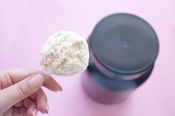 heap of whey protein powder with plastic spoon on a female hand and a can of protein in the background on pink background