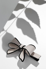 Subject shot of a cambered hair clip made of beige glazy material in the form of a bow-knot with a black stripe. The image is made in the studio on the white surface with shadows of the leaves. © RedUmbrella&Donkey