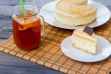Classic cheesecake cake on a dish, a piece of cake on a saucer, a spoon, tea in a mug, everything on a straw napkin on a dark wood tabletop