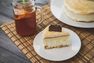 Classic cheesecake cake on a dish, a piece of cake on a saucer, a spoon, tea in a mug, everything on a straw napkin on a dark wood tabletop