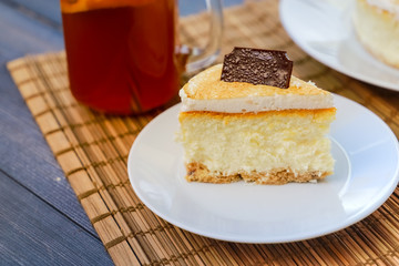 Classic cheesecake cake on a dish, a piece of cake on a saucer, a spoon, tea in a mug, everything on a straw napkin on a dark wood tabletop