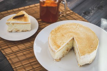 Classic cheesecake cake on a dish, a piece of cake on a saucer, a spoon, tea in a mug, everything on a straw napkin on a dark wood tabletop