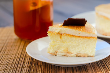 Classic cheesecake cake on a dish, a piece of cake on a saucer, a spoon, tea in a mug, everything on a straw napkin on a dark wood tabletop