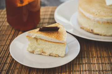 Classic cheesecake cake on a dish, a piece of cake on a saucer, a spoon, tea in a mug, everything on a straw napkin on a dark wood tabletop