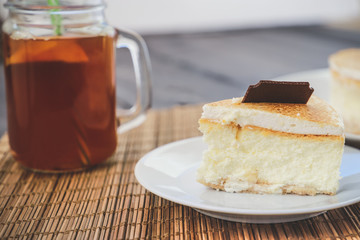 Classic cheesecake cake on a dish, a piece of cake on a saucer, a spoon, tea in a mug, everything on a straw napkin on a dark wood tabletop