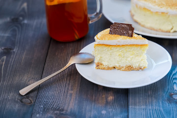 Classic cheesecake cake on a dish, a piece of cake on a saucer, a spoon, tea in a mug, everything on a straw napkin on a dark wood tabletop