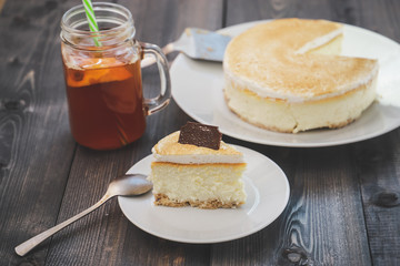Classic cheesecake cake on a dish, a piece of cake on a saucer, a spoon, tea in a mug, everything on a straw napkin on a dark wood tabletop