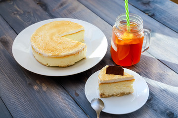 Classic cheesecake cake on a dish, a piece of cake on a saucer, a spoon, tea in a mug, everything on a straw napkin on a dark wood tabletop