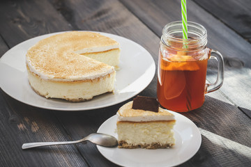 Classic cheesecake cake on a dish, a piece of cake on a saucer, a spoon, tea in a mug, everything on a straw napkin on a dark wood tabletop