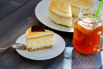 Classic cheesecake cake on a dish, a piece of cake on a saucer, a spoon, tea in a mug, everything on a straw napkin on a dark wood tabletop