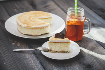 Classic cheesecake cake on a dish, a piece of cake on a saucer, a spoon, tea in a mug, everything on a straw napkin on a dark wood tabletop