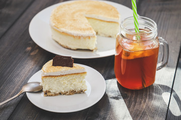 Classic cheesecake cake on a dish, a piece of cake on a saucer, a spoon, tea in a mug, everything on a straw napkin on a dark wood tabletop