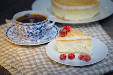 On the saucer is a piece of cheesecake cake and red berries, the whole cake on a white plate, next to it is a white tea pair with a blue pattern, everything is on a linen napkin.