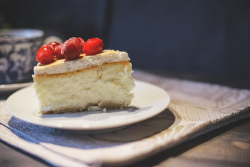 On a white saucer, a piece of cheesecake cake with red berries laid out on top, next is a white tea pair with a blue pattern.