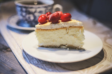 On a white saucer, a piece of cheesecake cake with red berries laid out on top, next is a white tea pair with a blue pattern.