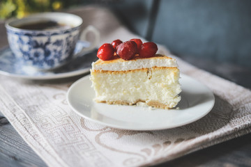 On a white saucer, a piece of cheesecake cake with red berries laid out on top, next is a white tea pair with a blue pattern.