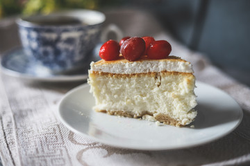 On a white saucer, a piece of cheesecake cake with red berries laid out on top, next is a white tea pair with a blue pattern.