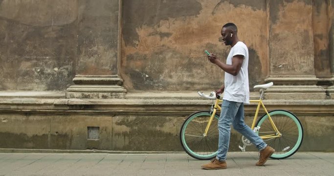 Afro American Young Man in Earphones Using his Modern Mobile Phone and Pushing Stylish Thin Tire Bike while Going along Old Building Wall.