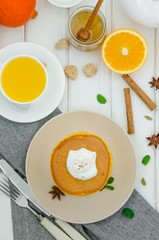 Stack of spicy pumpkin pancakes on a plate with honey and whipped cream on a white wooden background.  Rustic style. Fragrant autumn breakfast. Breakfast for Halloween.