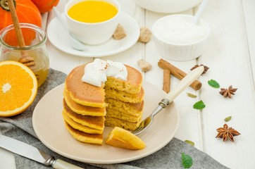 Stack of spicy pumpkin pancakes on a plate with honey and whipped cream on a white wooden background.  Rustic style. Fragrant autumn breakfast. Breakfast for Halloween.