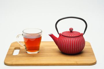 Cast-iron teapot on a wooden board and a glass cup with hot tea on a white background.