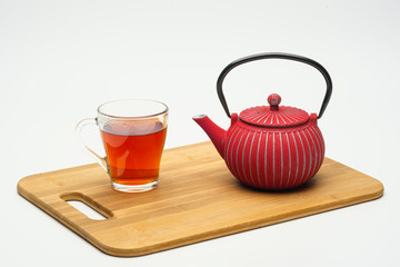Cast-iron teapot on a wooden board and a glass cup with hot tea on a white background.
