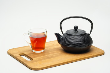 Cast-iron teapot on a wooden board and a glass cup with hot tea on a white background.