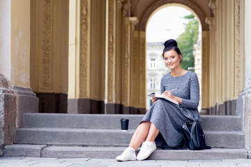 Student young woman is reading book while sitting on university stairs. Student adult girl sits on stairs while reads book near university.
