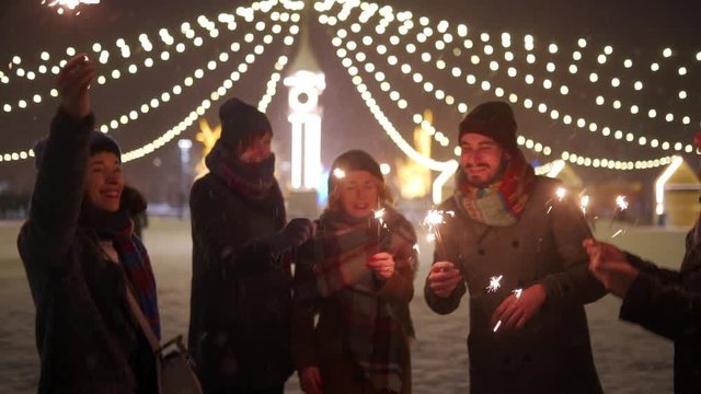 Cheerful friends having fun waving with sparklers in hands. Christmas eve, New Year Holidays or Birthday party. Happy people jump partying on winter night with snow and lamp garlands on background.