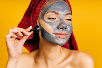 Charming young girl with a red towel on her head with a clay gray mask on her face and arrows holds the eyeliner cap on a yellow background and enjoys the mask