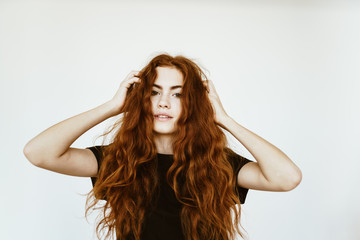 Lovely young girl with long curly red hair with freckles on her face in a black T-shirt on a white background holds her hands by her hair and poses