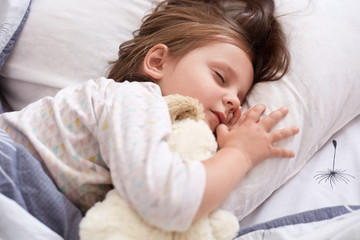 Indoor shot of pretty little girl hugging white soft dog toy while sleeping in bed at home, child spending morning time in cosy room, keeps eyes closed, having dark hair, desses white pajama.