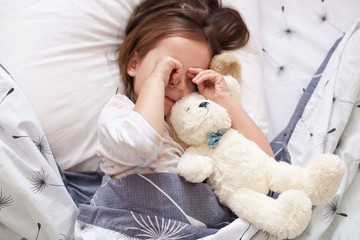 Portrait of lovely girl with white soft dog rubbing her eyes after sleep in morning, female kid lying on pillow in bed with her toy, does not want to stand up and go to kinder garten. Morning concept.