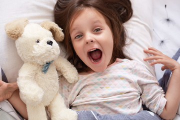 Indoor shot of cute darkhaired baby girl lying in bed, looking up with surprised look and opened mouth near white soft dog, beautiful kid dresses pajama looks astonished, relaxing in bed in morning.