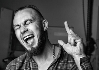 Cheerful excited man celebrating success. Bearded man in a checkered shirt rejoices against  background of the workshop. Shows symbol Rock-n-Roll. Men's Gift Concept. Hight contrast portrait