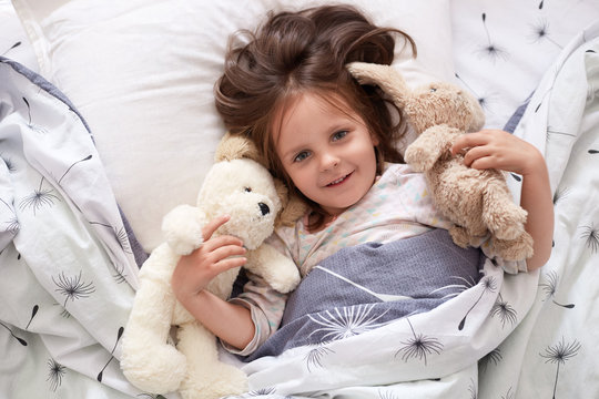 Image of girl lying with fluffy teddy bear and dog before giving them big hug, beautiful kid relaxing in bed with her toy, dark haired child wearing white pajama, charming kid expressing happyness.
