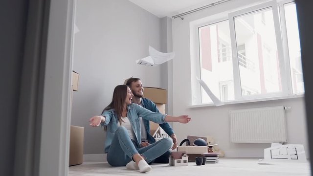 Happy joyful young couple in love sitting on the parquet and scattering design pictures in their new apartment,having fun together after relocation