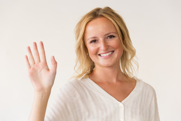 Obraz na płótnie Canvas Cheerful woman waving hand. Beautiful happy young blonde woman waving hand and smiling at camera on grey background. Gesture concept