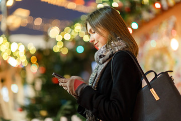 Photo side of woman with phone in hand on street in evening on blurred background with garland