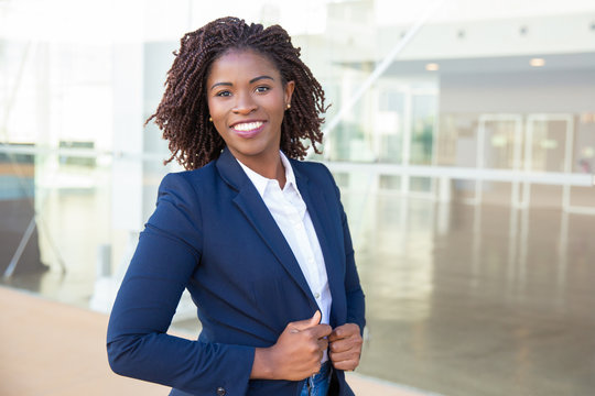 Happy confident professional posing near office building. Young African American business woman standing outside, adjusting formal jacket, looking at camera, smiling. Successful businesswoman concept