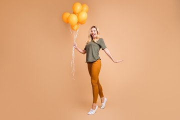 Full length profile photo of charming blond lady holding many bright orange air balloons came halloween party wear green t-shirt trousers isolated beige color background